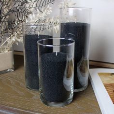 three glass vases filled with black sand on top of a wooden table next to a plant