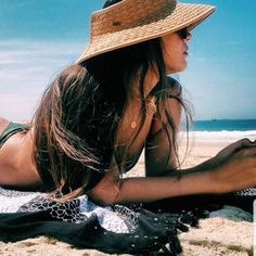 a woman laying on top of a beach next to the ocean wearing a straw hat