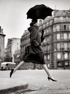 a woman is jumping in the air with an umbrella over her head while wearing a coat and heels