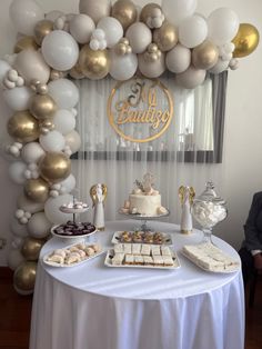 a table topped with lots of desserts under a balloon filled arch covered in white and gold balloons