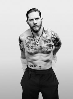 a man with tattoos on his chest standing in front of a white wall and looking at the camera