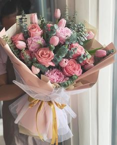 a woman holding a bouquet of pink roses and greenery in front of a window