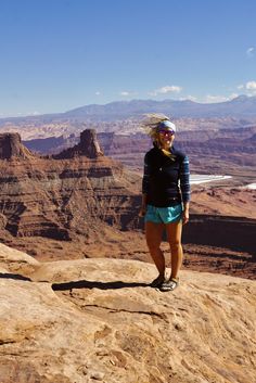 a woman standing on the edge of a cliff with mountains in the background and blue sky