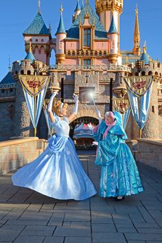 two women dressed in princess dresses dancing outside the castle at disneyland world, with their arms raised