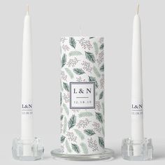 two white candles sitting next to each other on top of a glass container with holly leaves