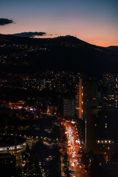 the city lights shine brightly at night in this view from atop a hill, with mountains in the background