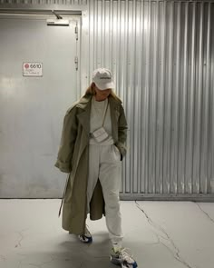 a man standing in front of a wall wearing a trench coat and white hat with his hands on his hips