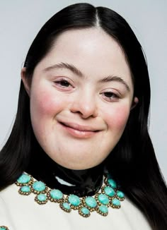 a woman with long black hair wearing a white top and turquoise jeweled collar smiling at the camera