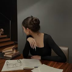 a woman sitting at a table with books and glasses on it, looking down into the distance