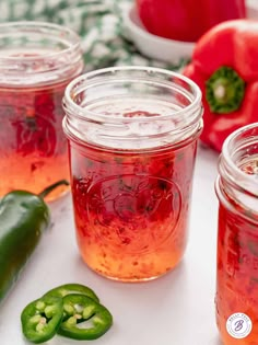 three jars filled with red liquid next to peppers and green peppers on a white surface