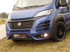 the front end of a blue van parked on top of a grass covered field with trees in the background