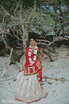 a woman in a red and white sari standing next to a tree on the beach