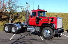 a red semi truck parked in a parking lot next to a tree and grass area