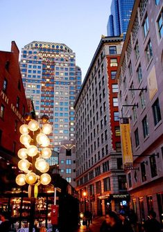 people are walking down the street at night in front of tall buildings with lights on them