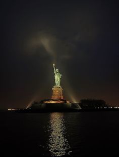 the statue of liberty is lit up at night with its lights on and reflecting in the water