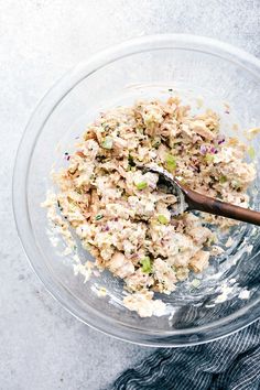 a glass bowl filled with tuna salad on top of a gray tablecloth next to a wooden spoon