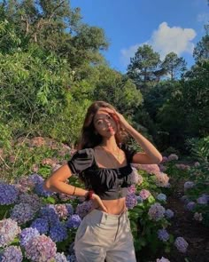 a woman standing in front of purple flowers with her hands on her head and eyes closed