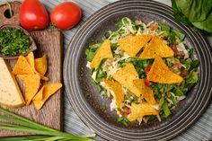 a salad with tortilla chips and tomatoes on a cutting board next to some greens