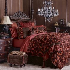 a bed with red and brown comforters in a room next to a dresser, chair and chandelier