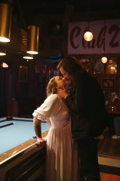 a man and woman are kissing in front of a pool table at a bar with lights hanging from the ceiling