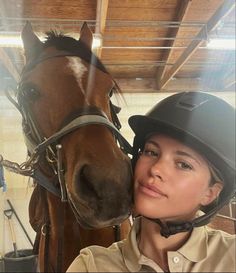 a woman wearing a helmet and standing next to a brown horse in a stable with it's head on the bridle