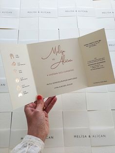 a person holding up a wedding card in front of a wall with white tiles on it