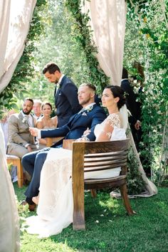 a bride and groom sitting on a wooden bench in front of an outdoor wedding ceremony