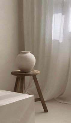 a white vase sitting on top of a wooden stand in front of a curtained window