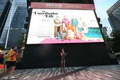 a woman standing in front of a large screen on the side of a building with people looking at it