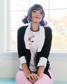 a woman with blue hair sitting in front of a window wearing pink pants and a black cardigan