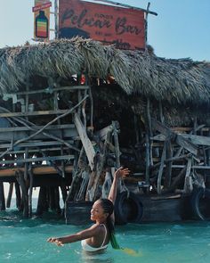 a woman standing in the water next to a building made out of wood and thatched roof