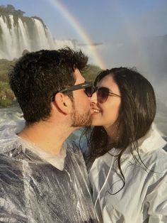 a man and woman are kissing in front of a waterfall with a rainbow behind them