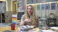 a woman is sitting at a table with some art supplies in front of her and pointing to the camera