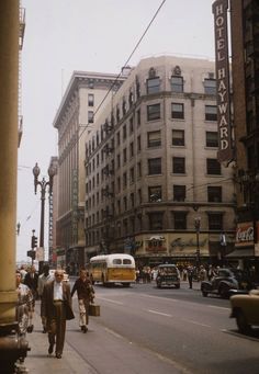 an old photo of people walking on the sidewalk in front of buildings and cars driving down the street