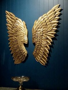 two gold angel wings on a blue wall next to a silver bowl and candle holder