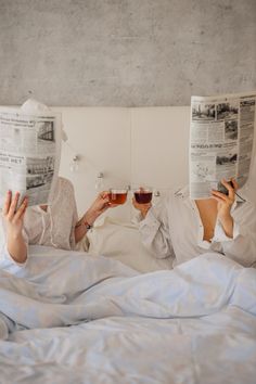 two women laying in bed reading newspapers and drinking wine