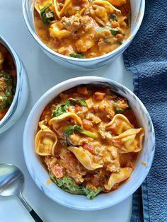 three bowls of pasta and spinach soup on a white surface with spoons next to it