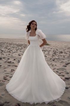 a woman is standing on the beach wearing a white wedding dress with sheer shoulders and sleeves