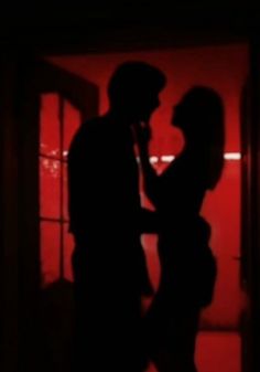 a man and woman standing next to each other in front of a red wall at night