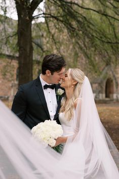 a bride and groom kissing in front of trees with their veil blowing in the wind