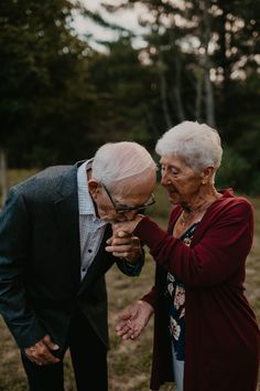 an older man and woman standing next to each other in a field with trees behind them