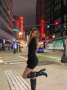 a woman in black dress and boots standing on street corner at night with traffic lights