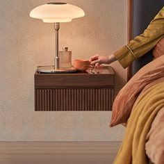 a woman sitting in bed next to a table with a cup on it and a lamp