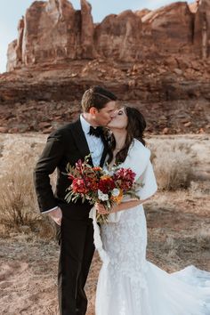 a bride and groom kissing in front of the desert landscape at their wedding day with red flowers