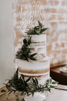 a three tiered cake with greenery on top sits on a table in front of a brick wall