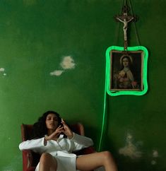 a woman sitting in a chair while holding a cell phone to her ear with a painting hanging on the wall behind her