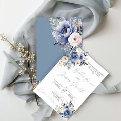 a wedding card with blue flowers and greenery on it next to a gray scarf