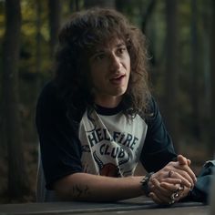 a woman with long hair sitting at a table talking to another person in the woods