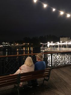 two people sitting on a bench looking out over the water at night with lights strung overhead