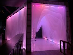 an empty room with white curtains and purple lights on the walls, in front of a staircase